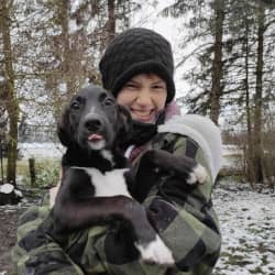 Benita Schmedemann lacht mit Wollmütze im leichten Schnee und hält einen schwarz-weißen Welpen im Arm