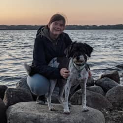 Xenia Vieth hockt in der Abenddämmerung auf Ufersteinen am Wasser, neben ihr ihre schwarz-weiße Hündin